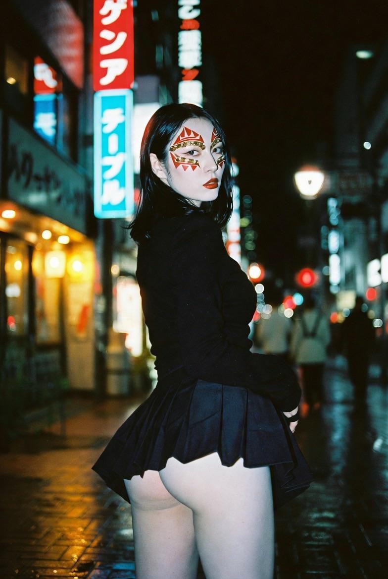 A woman with face paint and a short skirt stands on a wet street at night, looking over her shoulder.