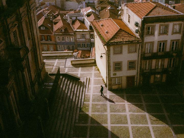 A person walks across a sunlit cobblestone square surrounded by buildings with red tile roofs.