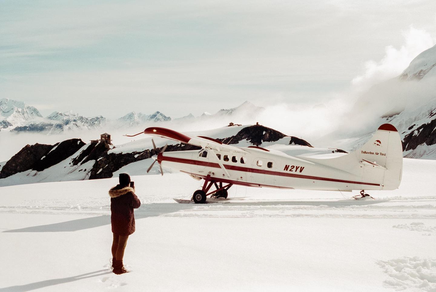 A person stands in a snowy landscape, photographing a small plane with mountains and clouds in the background.