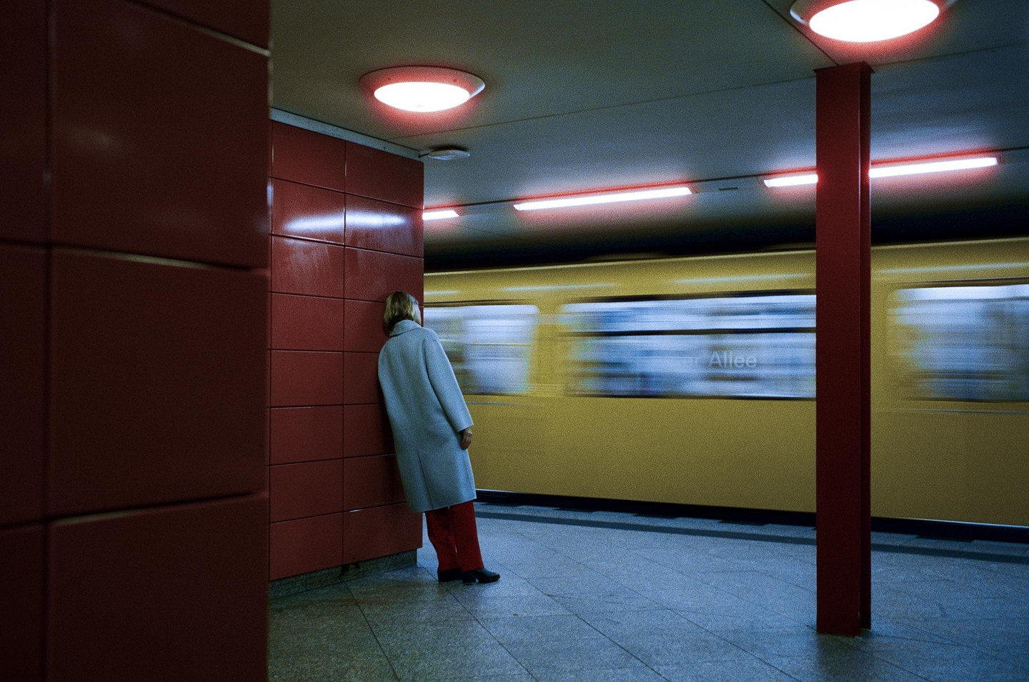 A woman leans against a red wall in a subway station as a yellow train passes by.