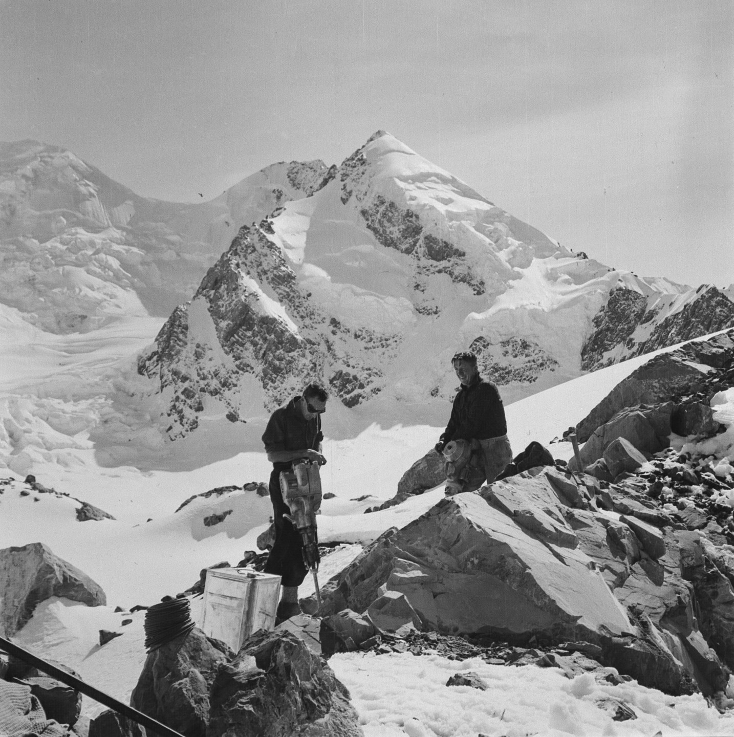Two men are working on a snowy mountain with a large snow-covered peak in the background.
