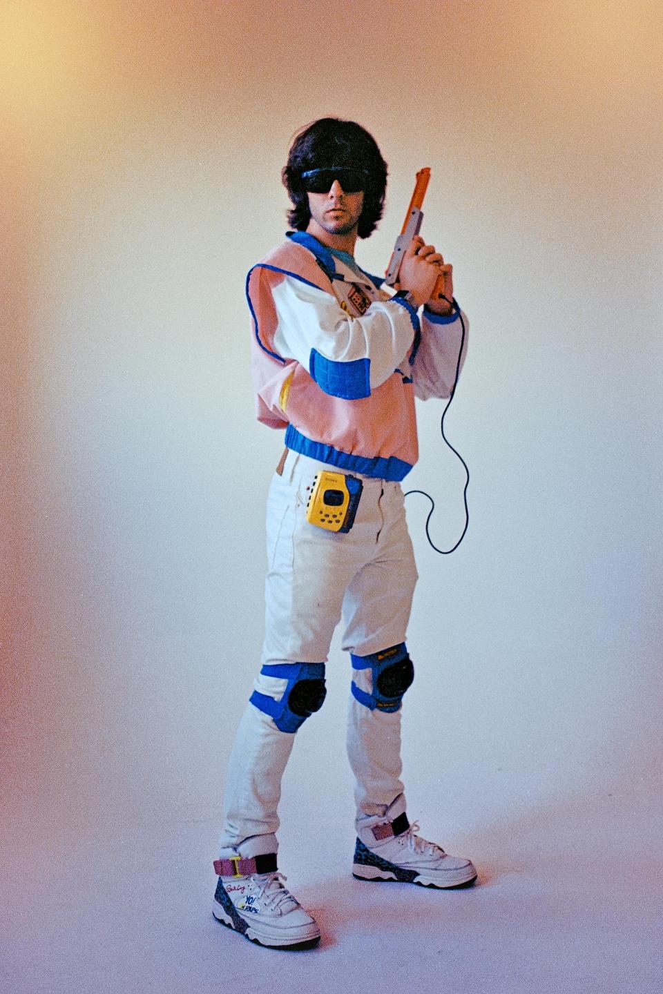 A man wearing sunglasses, a colorful jacket, white pants, and knee pads holds a toy gun and stands against a white background.