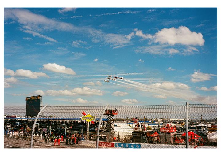 Six fighter jets fly in formation, leaving smoke trails in a blue sky with clouds above a racetrack.