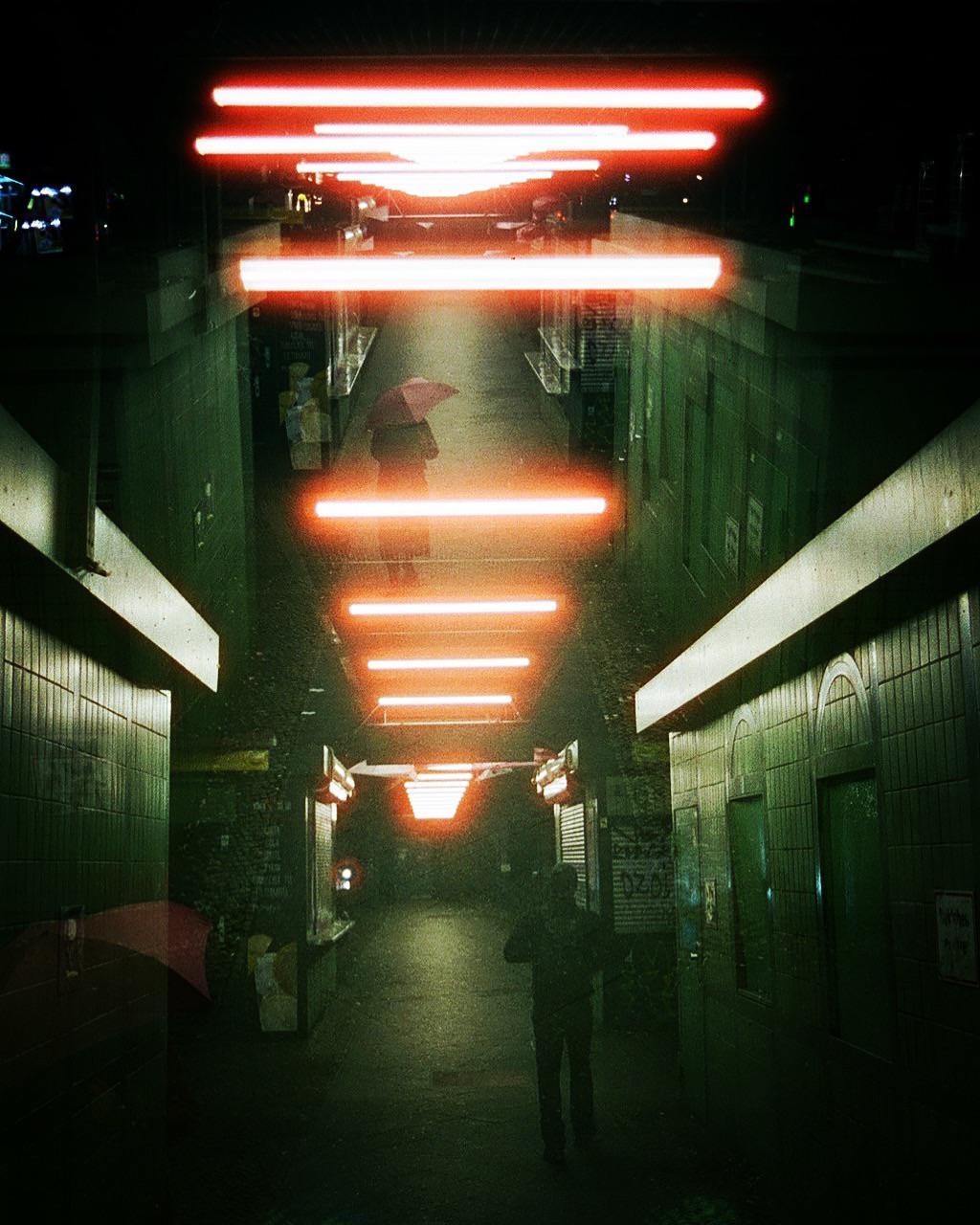 A person with an umbrella walks down a dark, tiled underpass illuminated by bright orange neon lights.