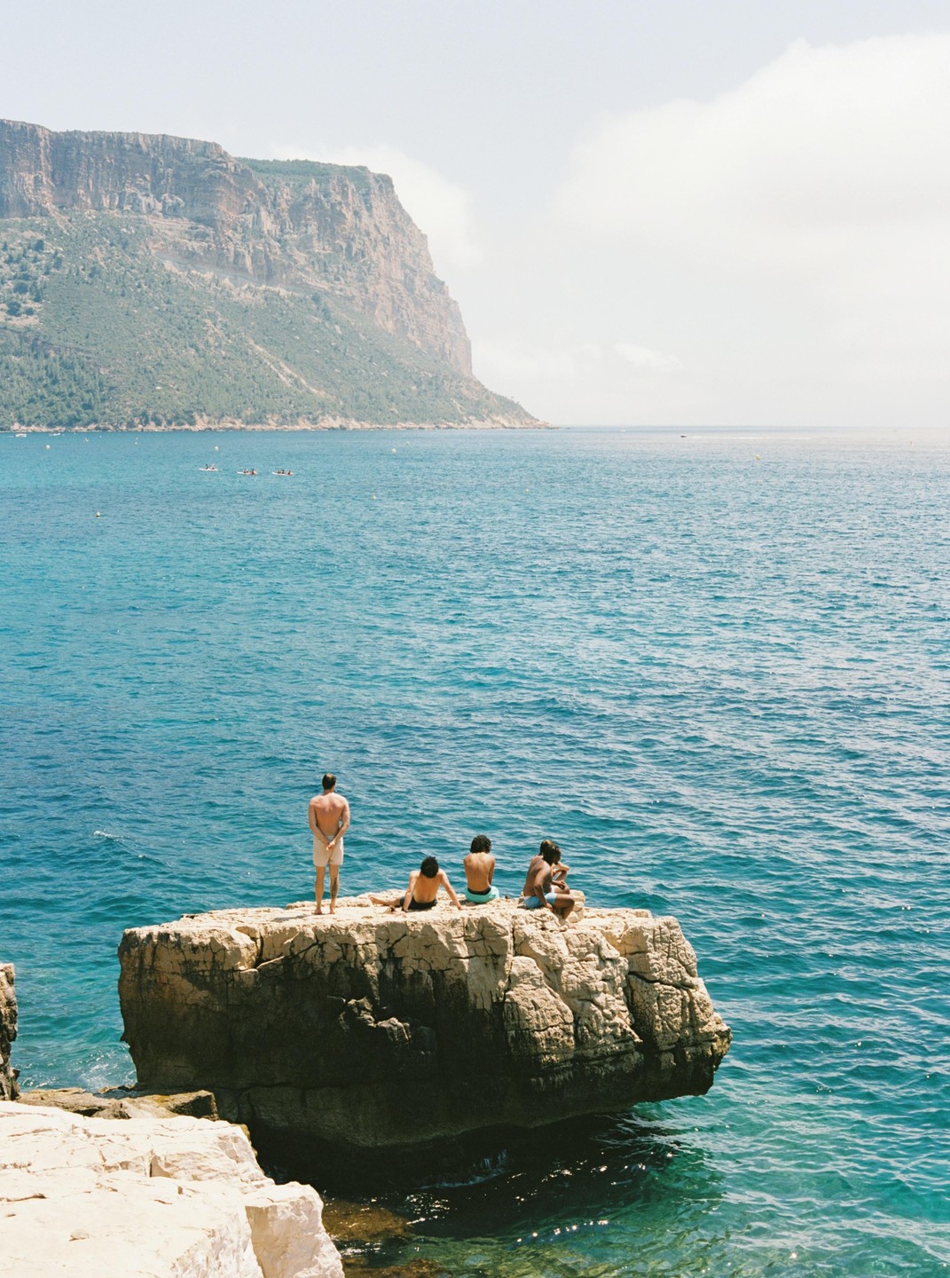 Four men are on a rock in the ocean with a large cliff in the background.