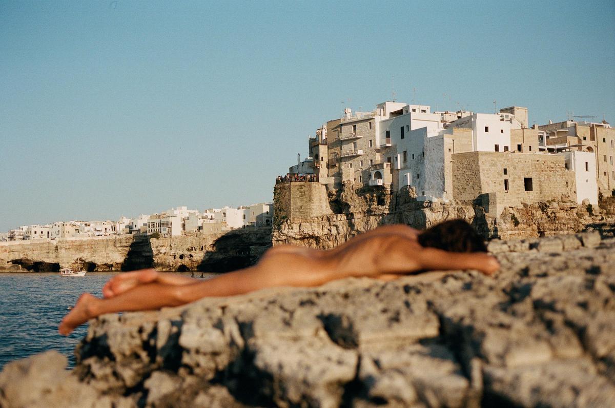 A nude woman lies on a rocky cliff overlooking the sea and a town built into the cliffs.