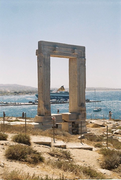 A large stone archway stands on a rocky outcrop overlooking a harbor with a ferry and sailboats.