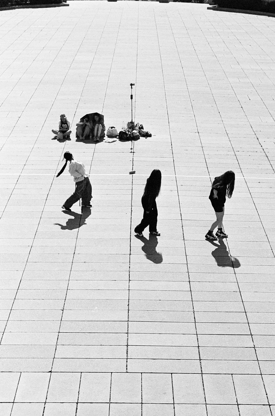 Three women walk across a large tiled plaza while three other people sit in the background.