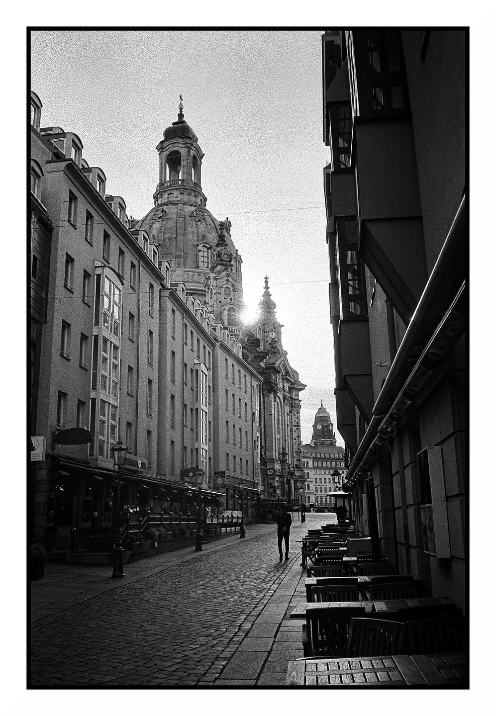 A man walks down a cobblestone street lined with buildings and outdoor seating, with a large domed building in the background.