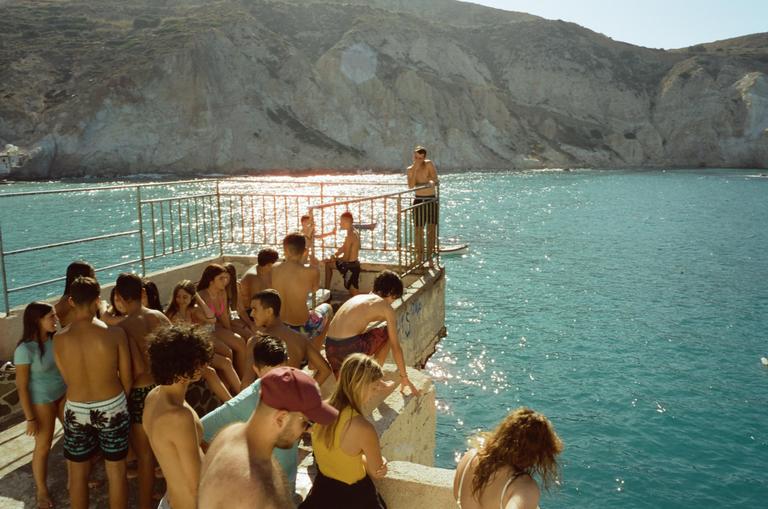 A group of young people gather on a concrete pier overlooking a bright blue sea with a rocky mountain in the background.