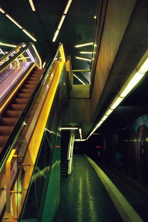 An escalator leads down to a dark subway platform illuminated by fluorescent lights.