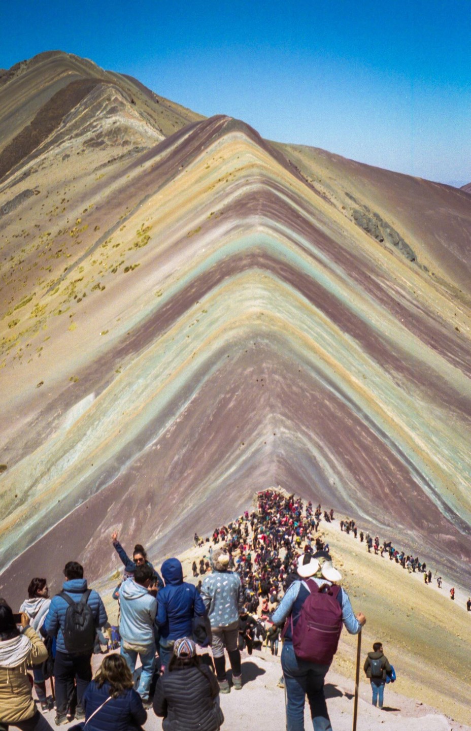A large crowd of people walk along a narrow path on a colorful mountain under a clear blue sky.