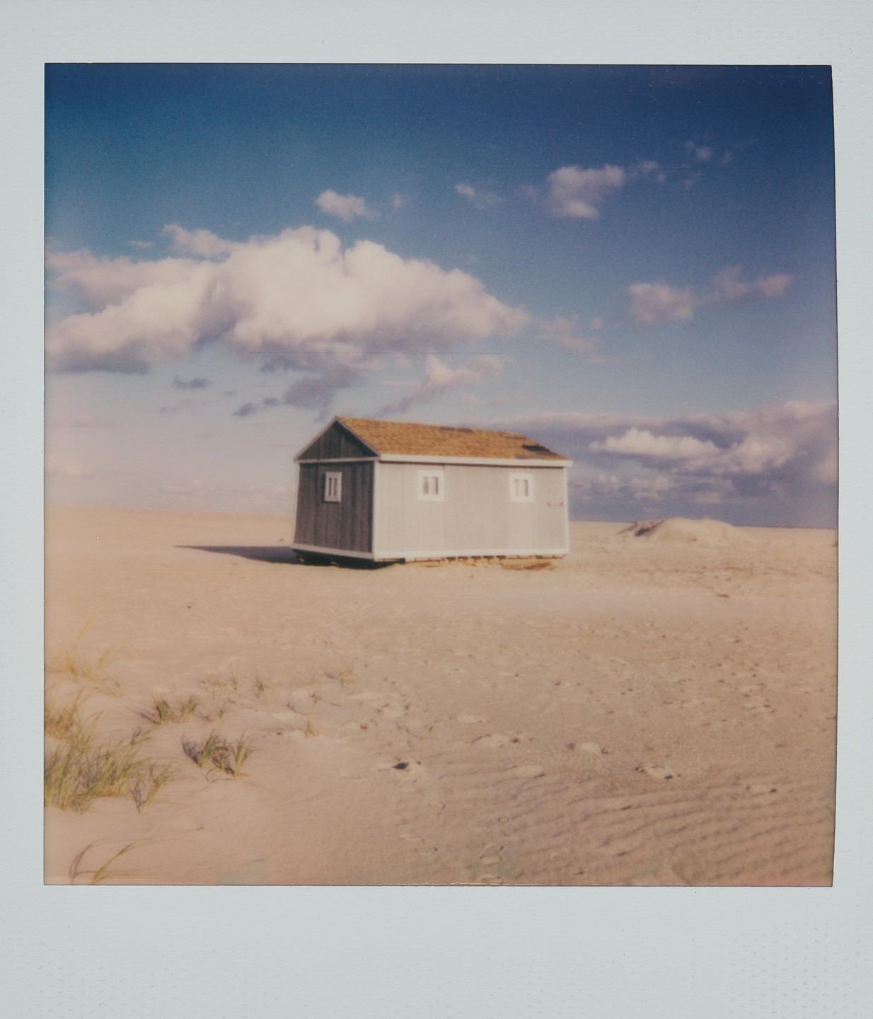 A small, light blue shack with a brown roof stands on a sandy beach under a blue sky with white clouds.
