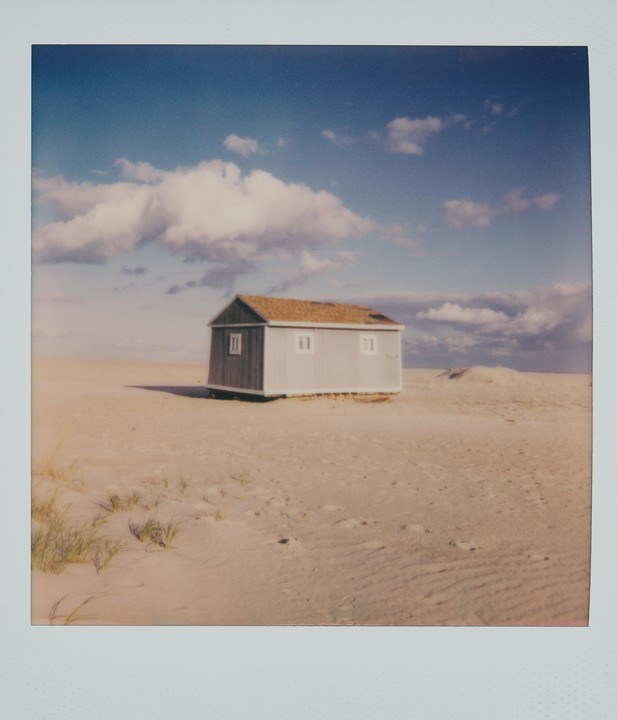 A small, light blue shack with a brown roof stands on a sandy beach under a blue sky with white clouds.