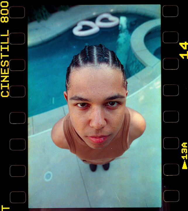 A man with braided hair and a tank top looks up at the camera from beside a swimming pool.