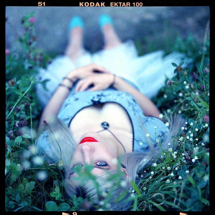 A woman with red lipstick and blue eyes lies in green grass and looks up at the viewer.