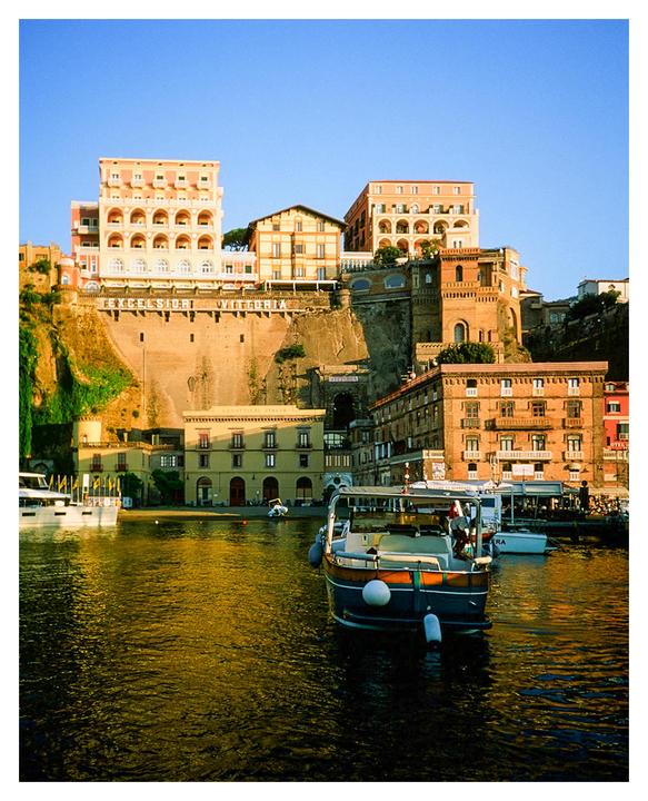 A boat is moored in a harbor with buildings on a cliff in the background.