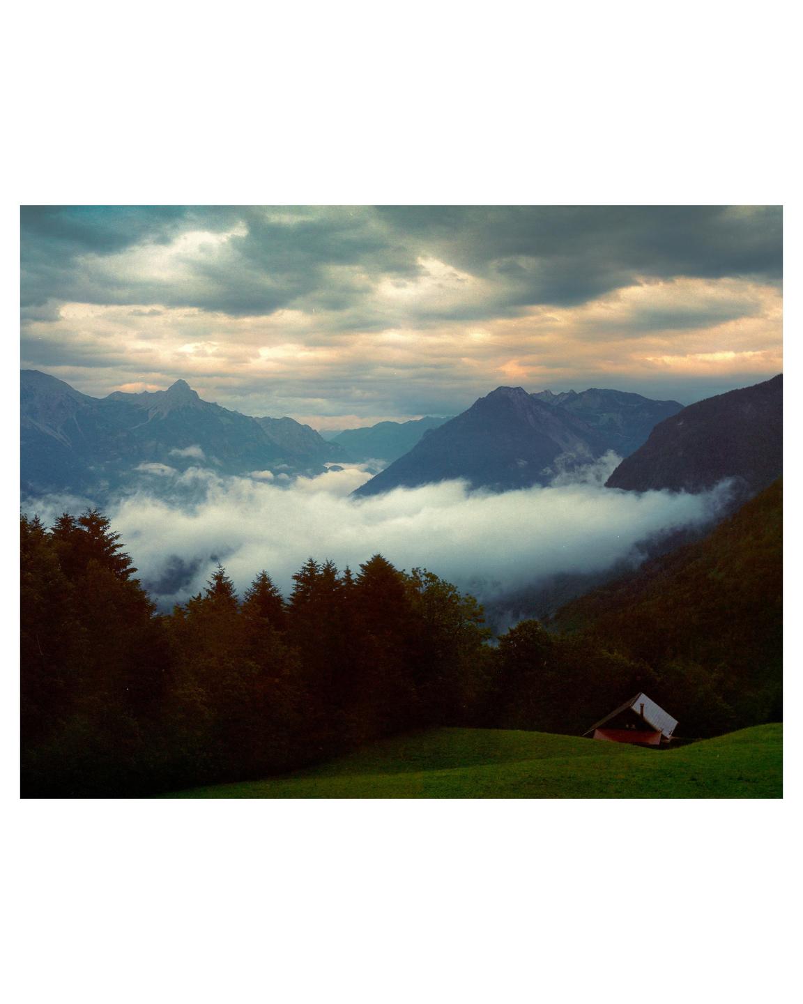 A small house sits on a green hillside overlooking a valley filled with fog and surrounded by mountains under a cloudy sky.
