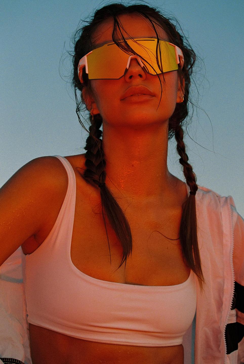 A woman with braided hair and mirrored sunglasses looks up against a blue sky.