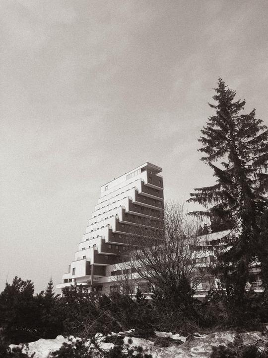A tall, stepped building stands among trees and snow under a bright sky.