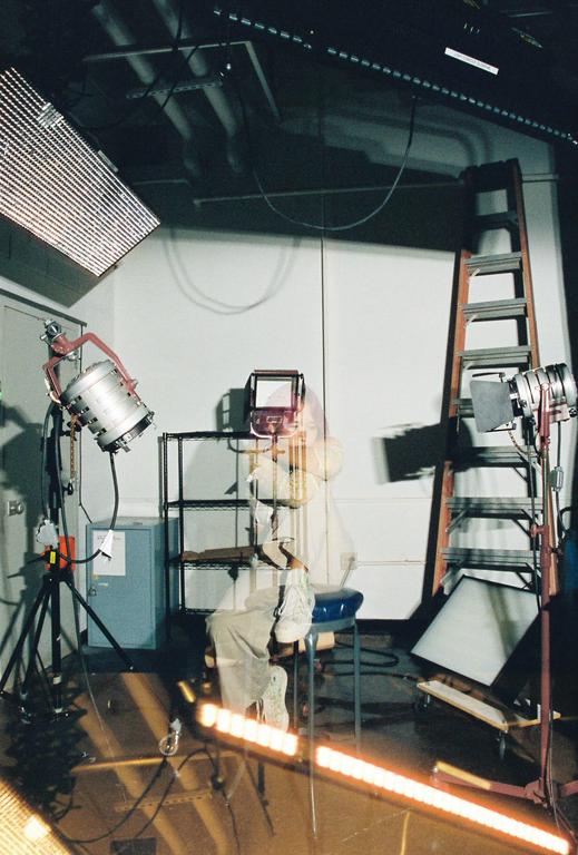 A person sits on a stool in a studio surrounded by various lighting equipment and a ladder.