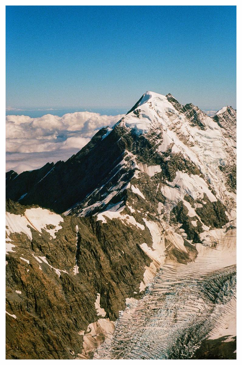 A snow-capped mountain peak rises above a glacier and clouds under a clear blue sky.