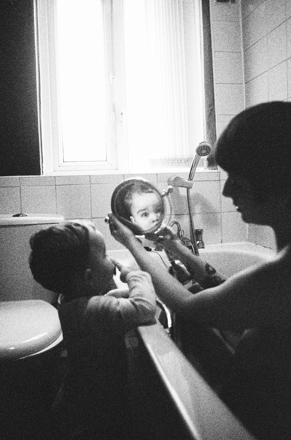 A child looks at their reflection in a handheld mirror while sitting in a bathtub with an adult.