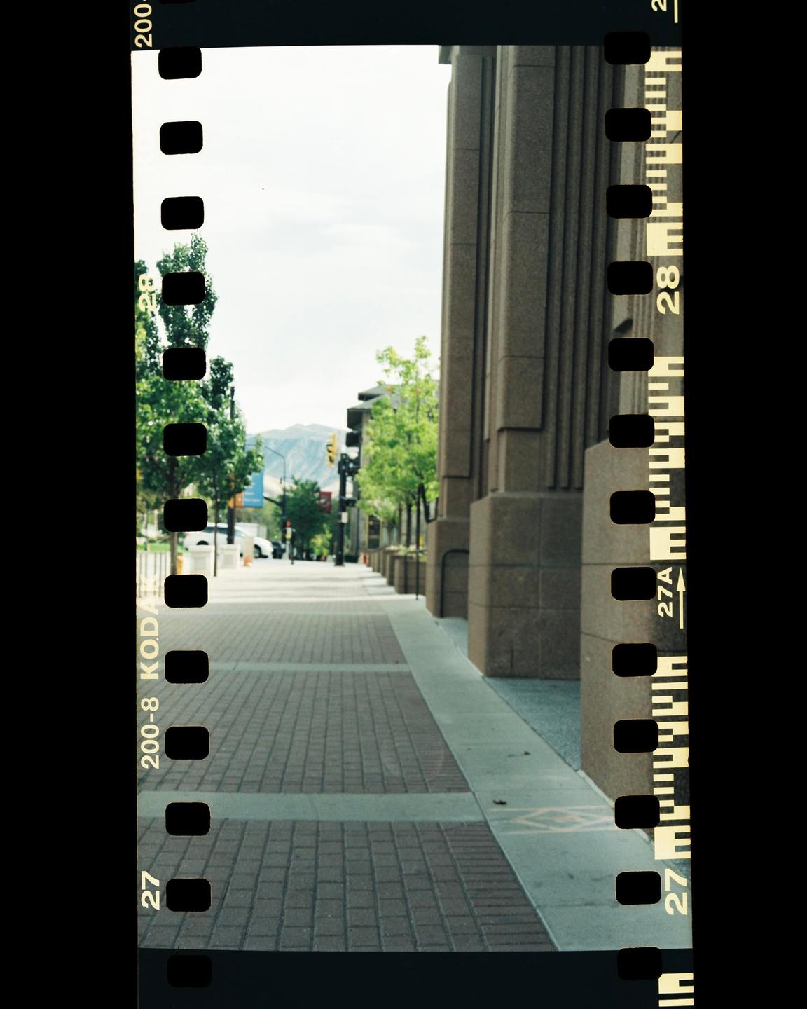 A wide sidewalk with brick pavers and a concrete border runs alongside a building with large columns and a street with trees and mountains in the distance.