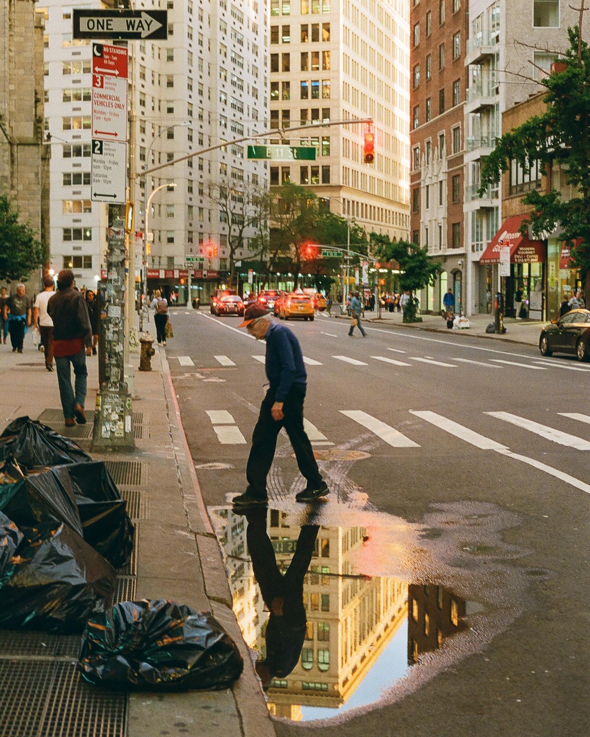 A man walks past a puddle reflecting a building on a city street with traffic and pedestrians.