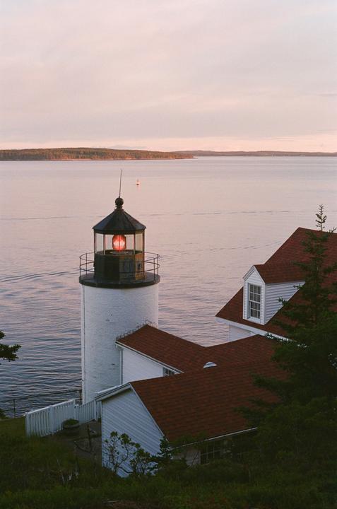 A white lighthouse with a red light is visible next to a building with a red roof, overlooking a body of water with a distant tree-lined shore.