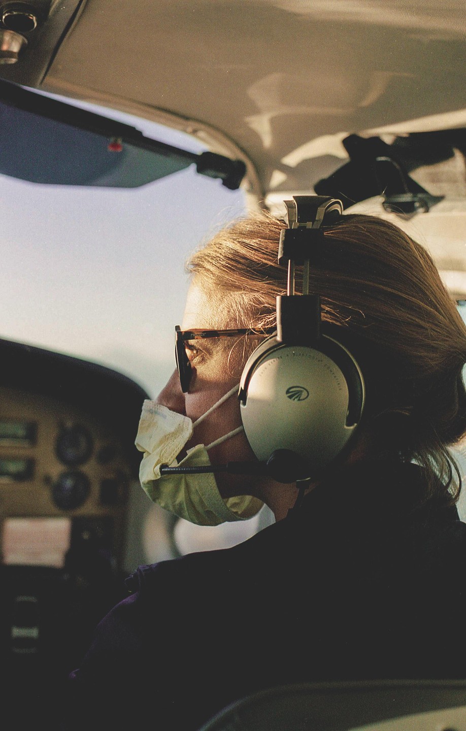 A woman wearing a face mask and headphones looks out the window of an airplane.