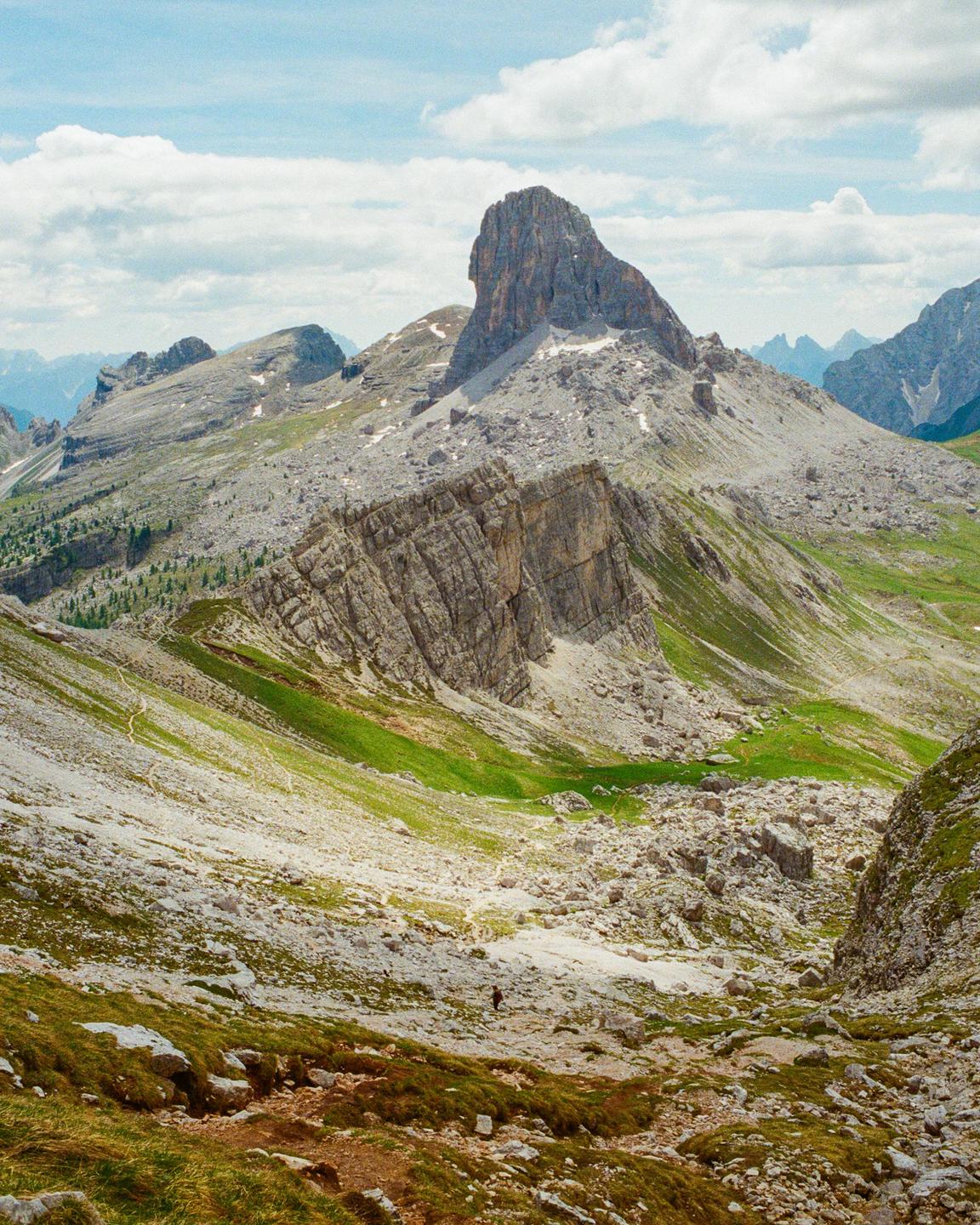 A person walks along a path through a rocky mountain landscape with patches of green grass and a prominent peak in the background.