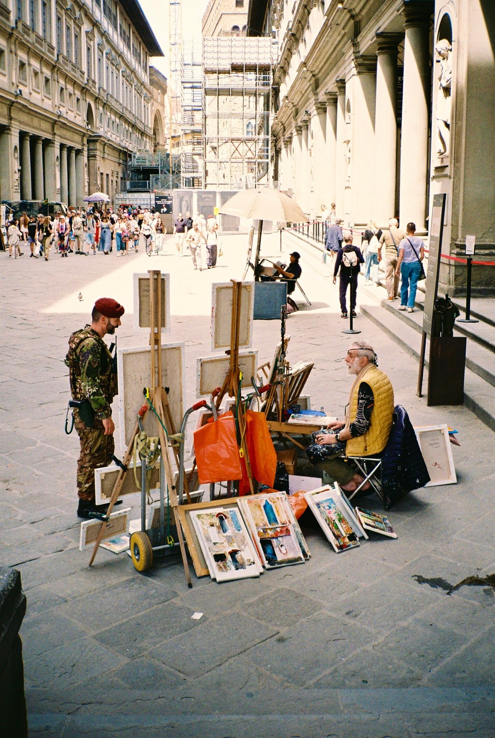 A street artist sits with his paintings and art supplies while a man in military fatigues stands nearby in a city square.