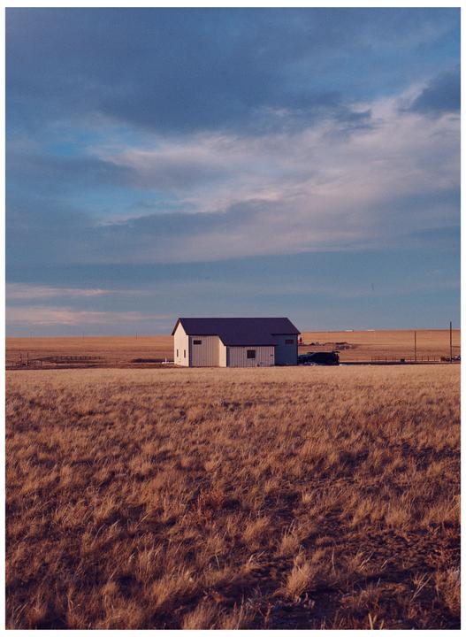 A white barn with a dark roof stands in a field of dry grass under a cloudy sky.