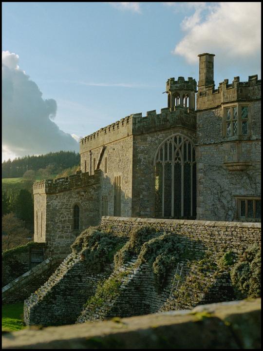 A stone castle with large windows and battlements stands on a hill, with a forest and cloudy sky in the background.