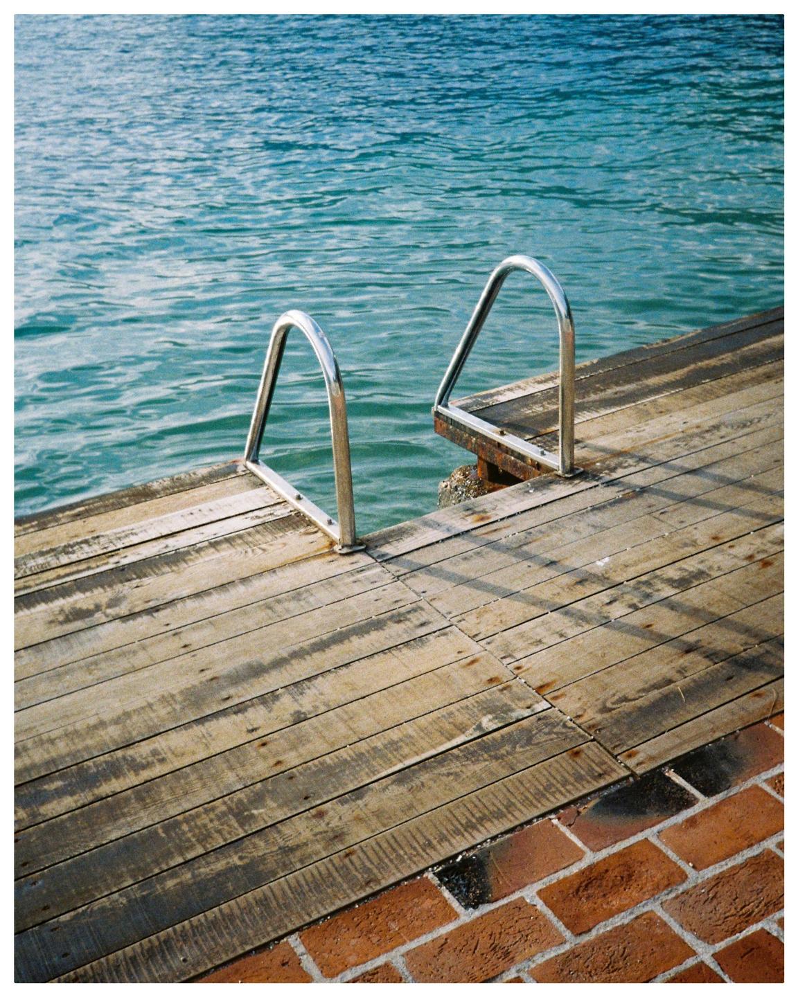 A metal ladder extends from a wooden dock into the blue water next to a brick path.