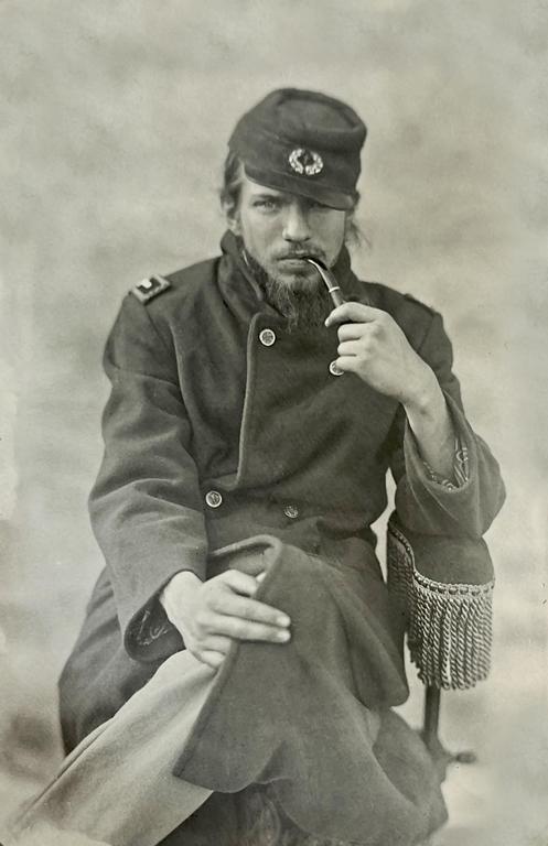 A man in a military uniform with a beard and long hair sits holding a pipe in his mouth.
