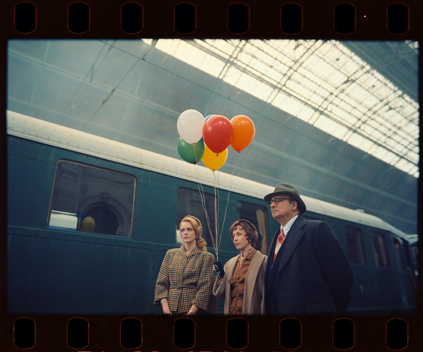A man and two women stand on a train platform with balloons.