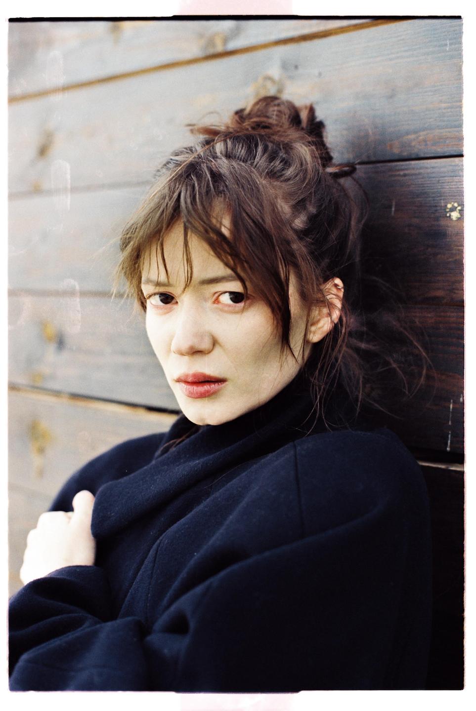 A woman with her hair in a bun and wearing a dark coat looks directly at the viewer.