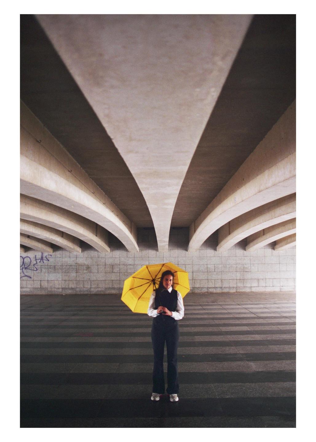 A woman stands under a yellow umbrella beneath a concrete overpass.