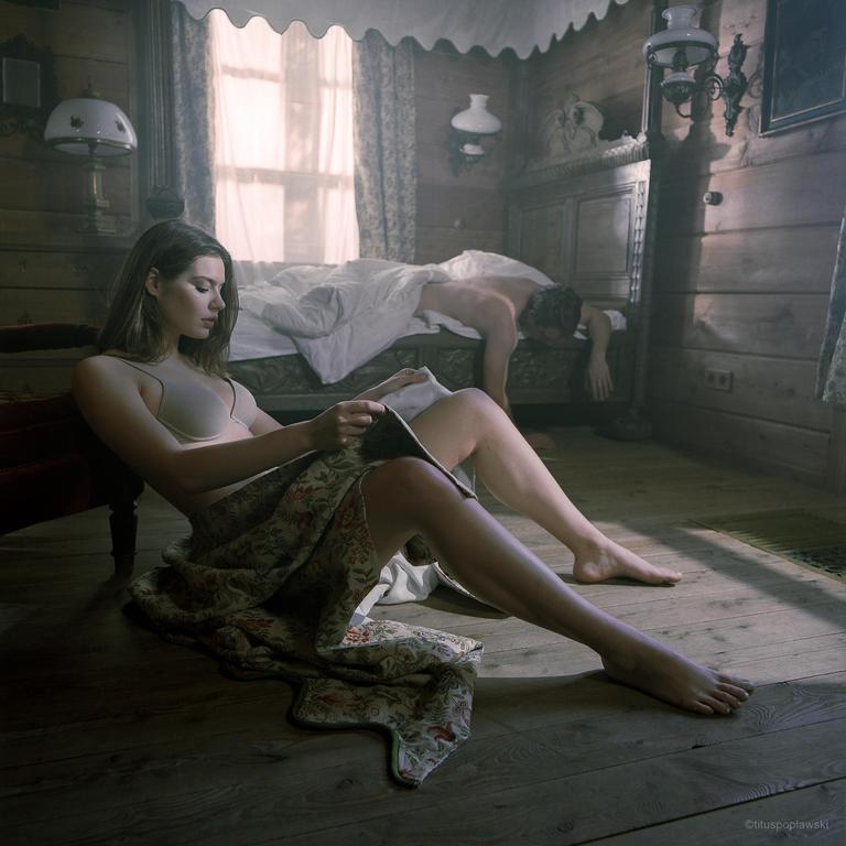A woman sits on the floor reading a book while a man sleeps in a bed behind her.