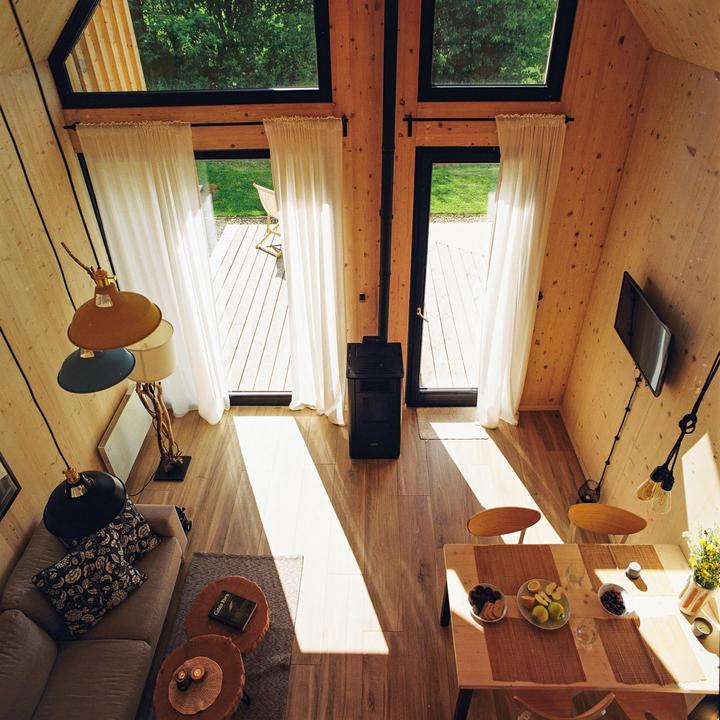 Sunlight streams into a modern cabin with a high ceiling, revealing a living area, dining table, and a view of a deck and trees outside.