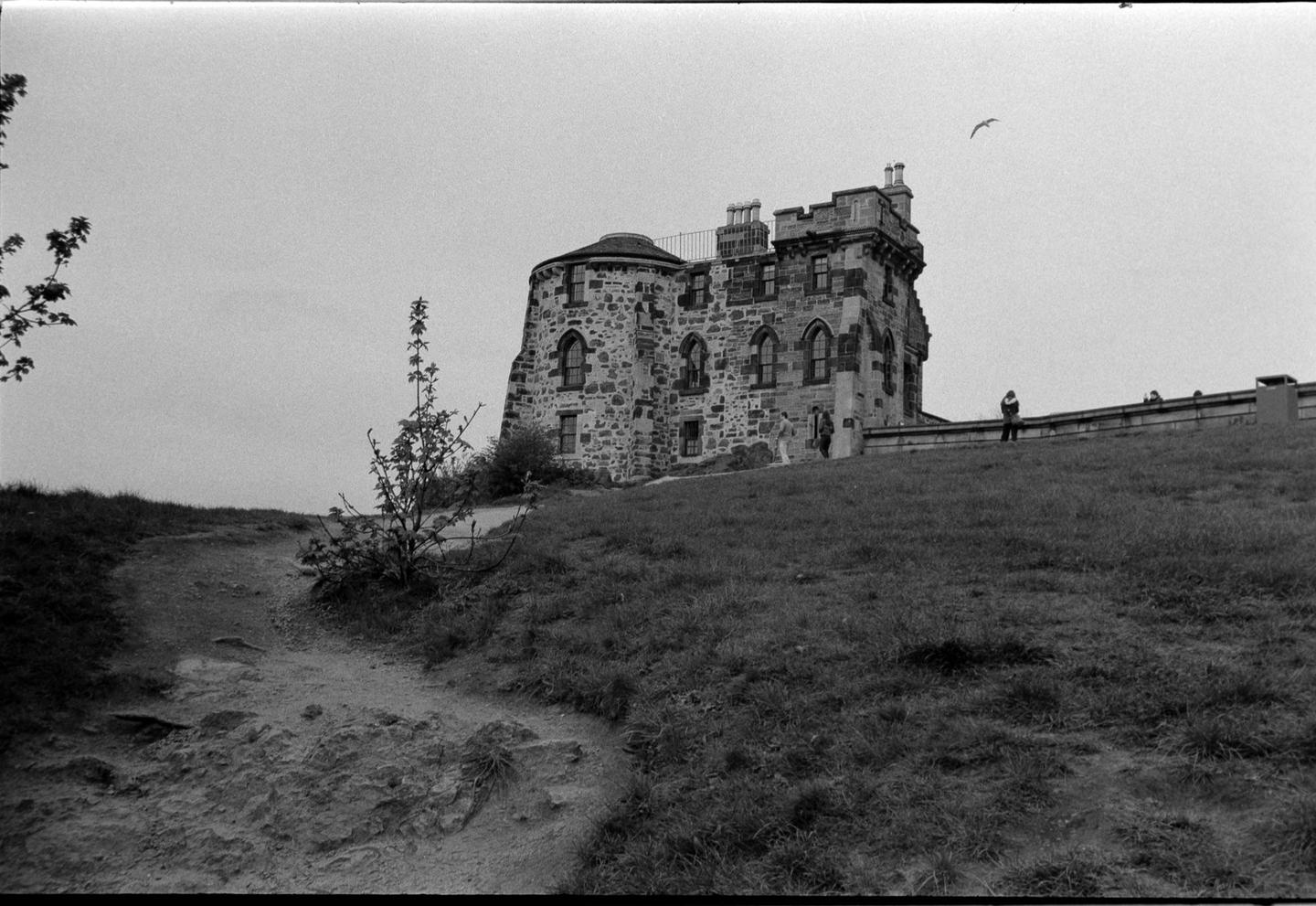 A stone building with turrets stands on a grassy hill with a dirt path in the foreground.