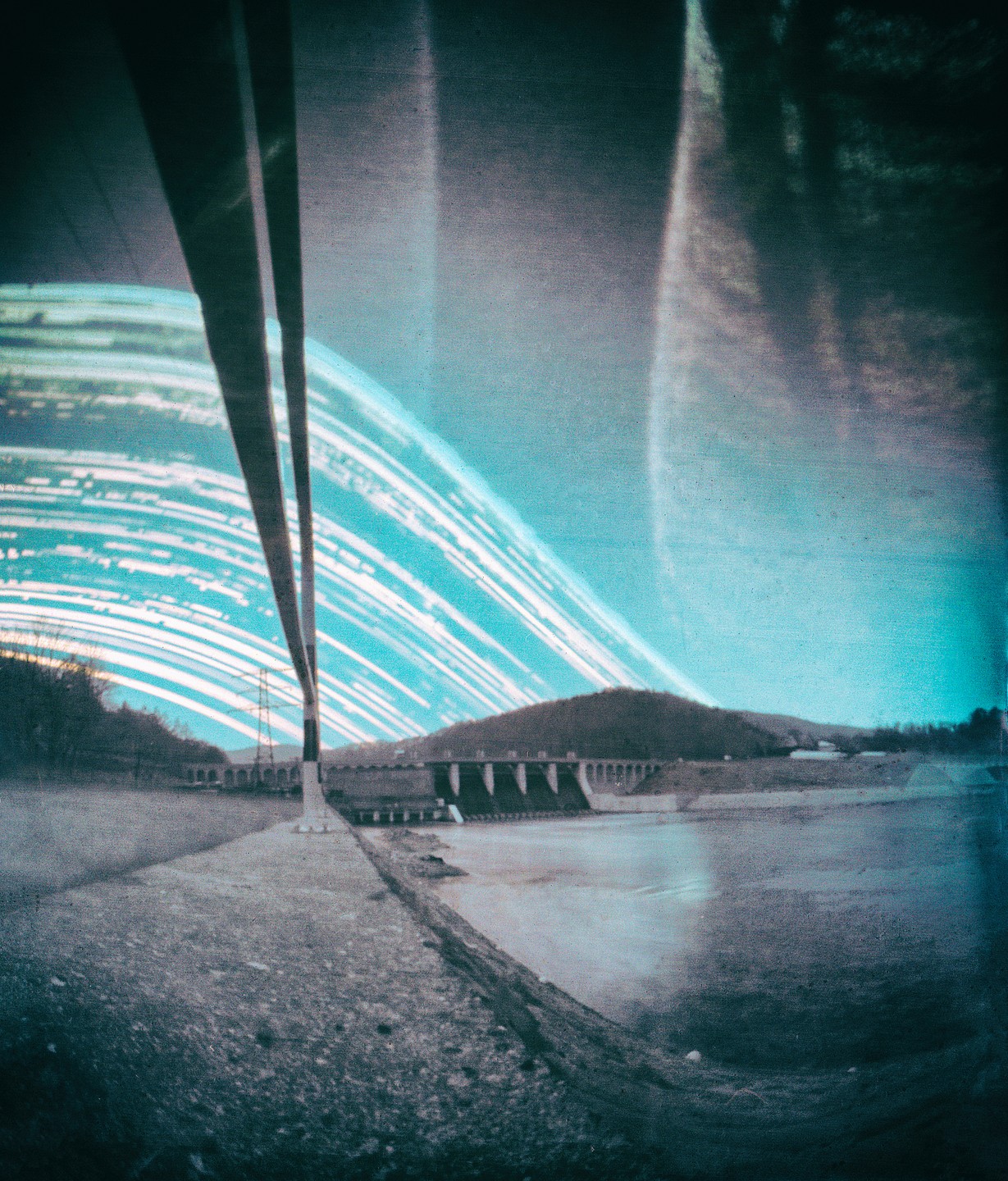 A long exposure photograph shows star trails over a dam and a body of water, with two dark vertical lines in the foreground.