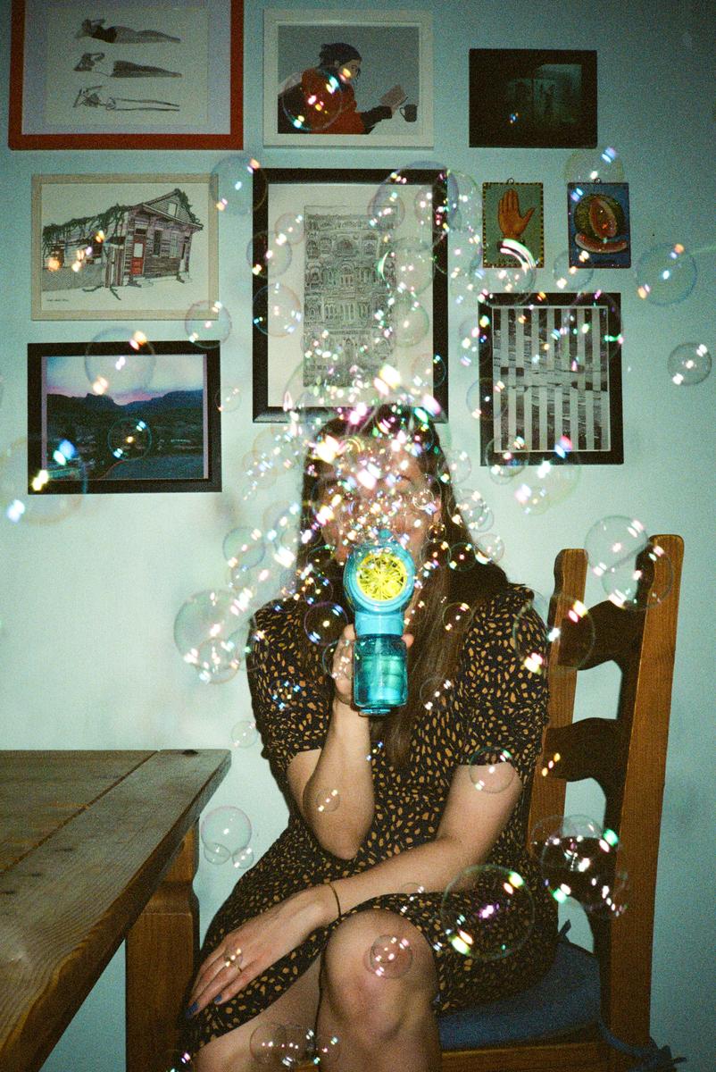A woman sits at a table and blows bubbles from a toy bubble gun.