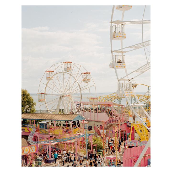 Two ferris wheels stand over a colorful amusement park filled with people and rides next to a body of water.