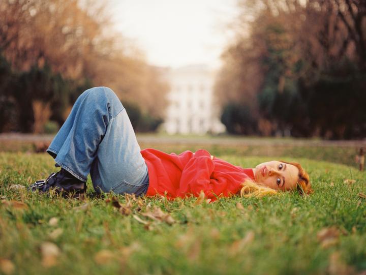 A woman in a red top and blue jeans lies on green grass with autumn trees and a building in the background.