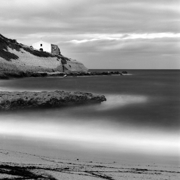 A small church sits atop a cliff overlooking a beach and the ocean under a cloudy sky.
