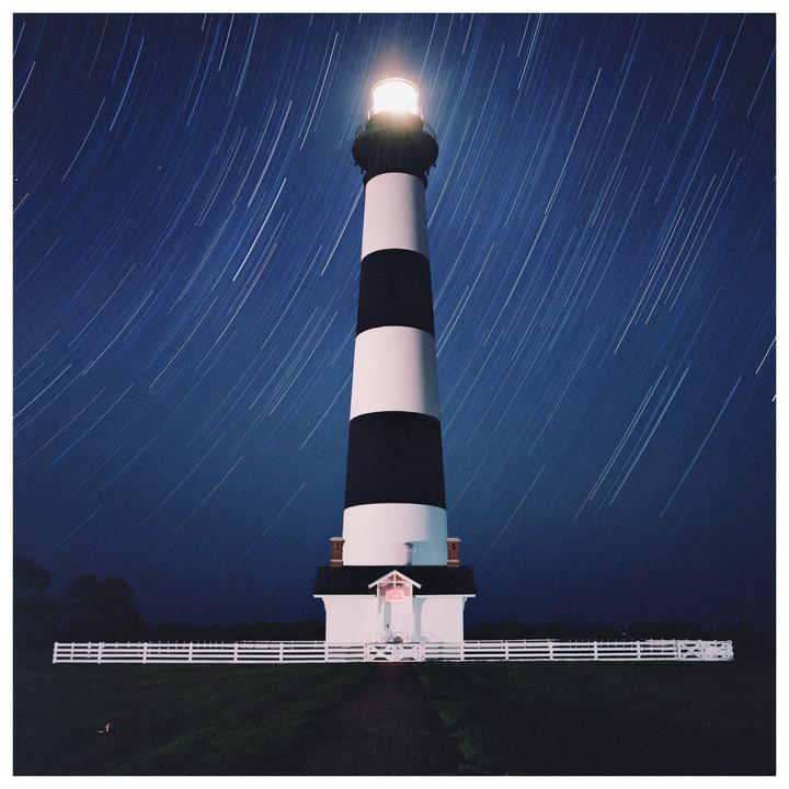 A black and white striped lighthouse stands tall against a dark blue night sky with star trails.