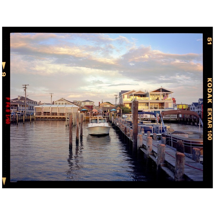 A boat is docked next to a pier with houses and other boats in the background under a cloudy sky.